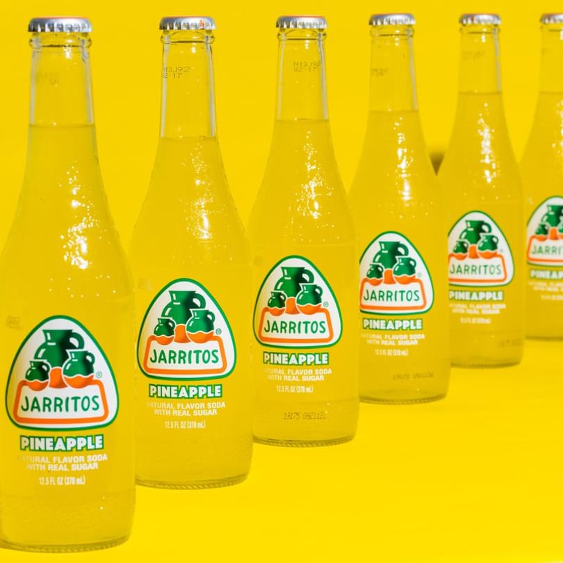 A row of Jarritos Pineapple soda bottles with yellow liquid and labels on a yellow background.