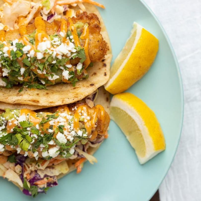 Two fish tacos with slaw, guacamole, cheese, cilantro, and sauce, served with lemon wedges.