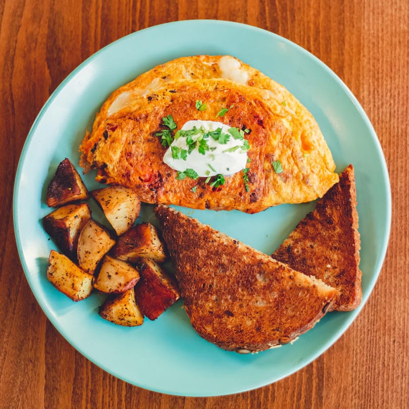Omelet topped with sour cream and herbs, served with roasted potatoes and toasted bread on a blue plate.
