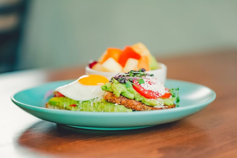 Avocado toast with a fried egg, tomatoes, and microgreens, served with a side of fruit.