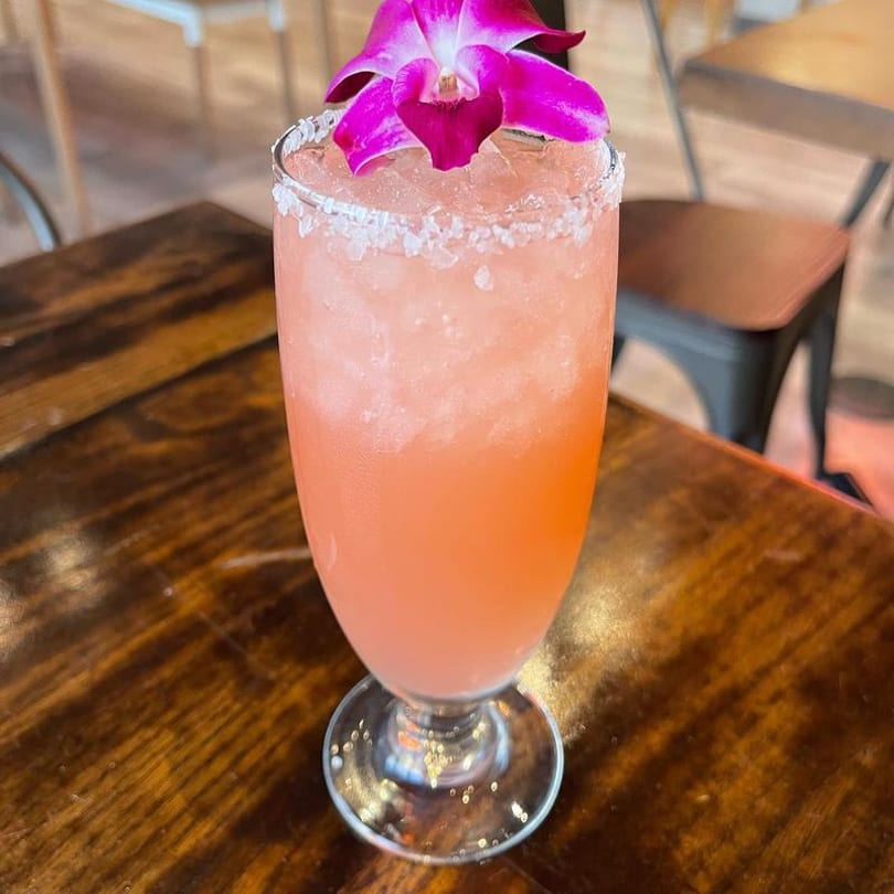 Pink cocktail with a salted rim and a purple orchid garnish on a wooden table.