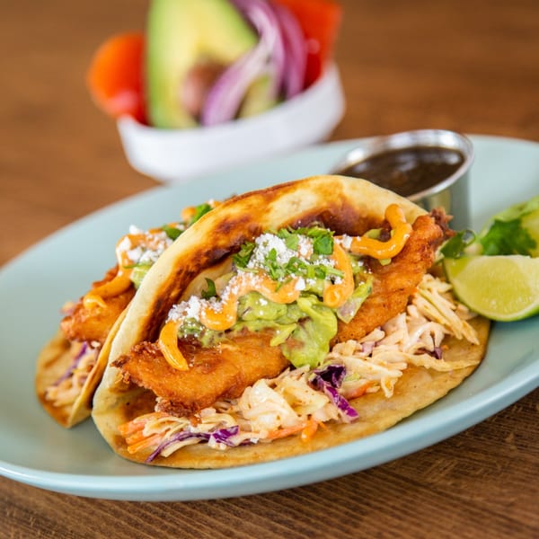 Two crispy fish tacos with slaw, guacamole, and sauce, served with lime wedges and a side of salsa.