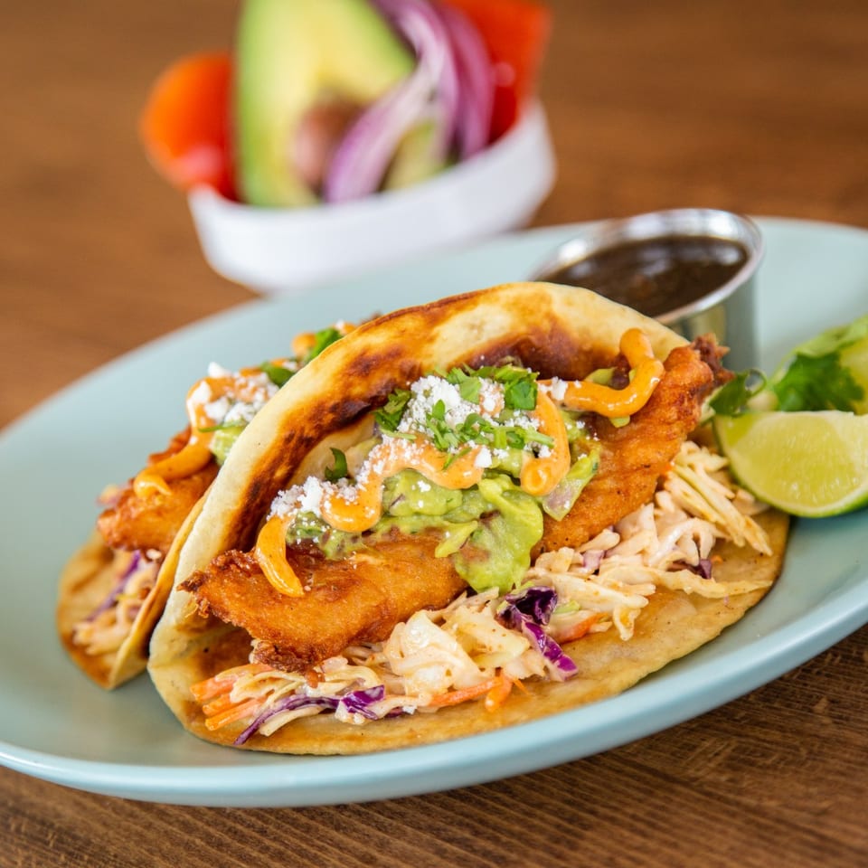 Two crispy fish tacos with slaw, guacamole, and sauce, served with lime wedges and a side of salsa.