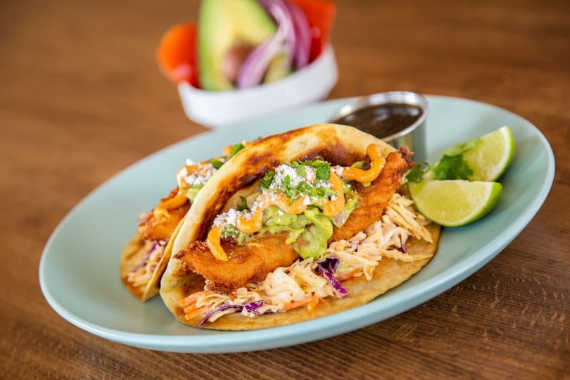 Two crispy fish tacos with slaw, guacamole, and sauce, served with lime wedges and a side of salsa.