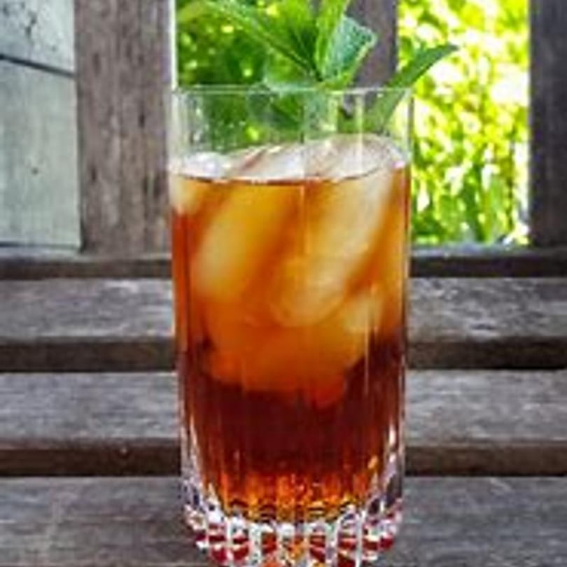 Iced tea with mint garnish in a tall glass on a wooden surface with greenery in the background.