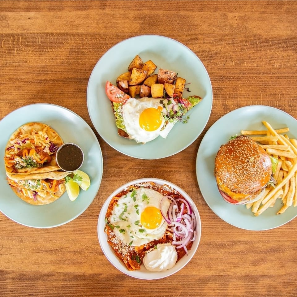 Four plates of food including tacos, avocado toast, chilaquiles, and a burger with fries.