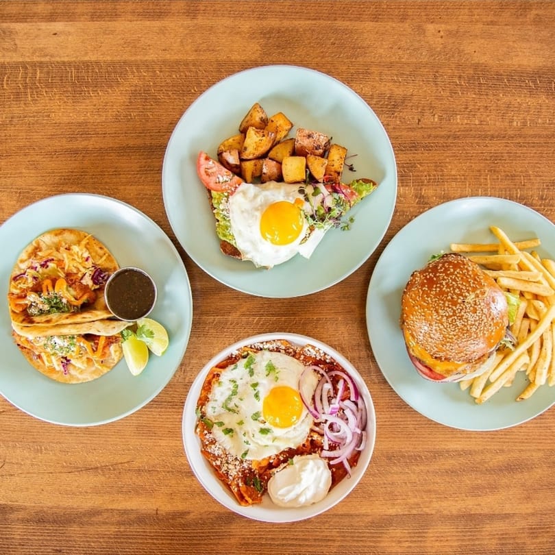 Four plates of food including tacos, avocado toast, chilaquiles, and a burger with fries.