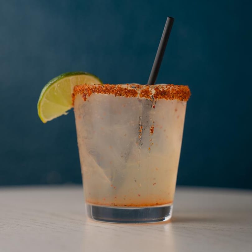 A chili-lime rimmed margarita with a lime wedge and black straw against a blue background.