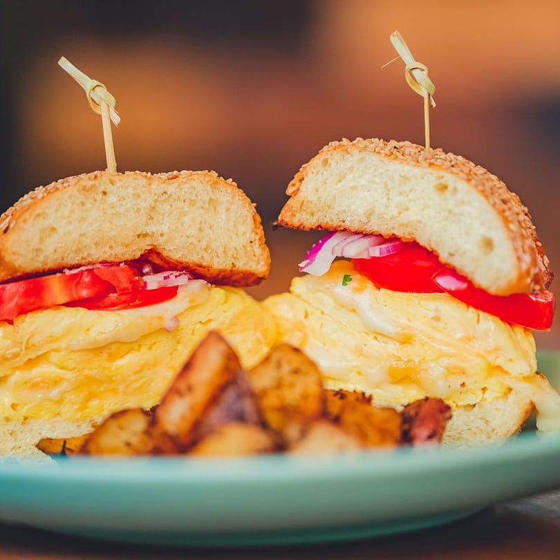 Two breakfast sandwiches with scrambled eggs, tomatoes, and onions, served with roasted potatoes.