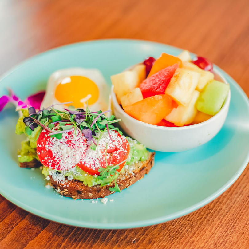 Avocado toast with tomatoes, microgreens, a fried egg, and a side of fruit salad.